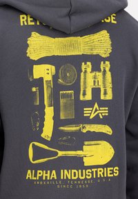 Tummanharmaa huppari, jossa on keltainen piirros erilaisista työkaluista ja varusteista, kuten lapio, veitsi ja kiikarit, sekä alapuolella brändinimi.