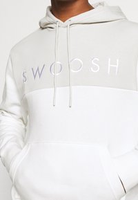 Huvtröja i vitt och ljusgrått. Har stort "SWOOSH"-logotyp i broderad text, känguruficka och snoddar.