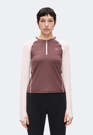 Frau mit kurzen dunklen Haaren, die ein braun-beiges langärmliges Zip-up-Oberteil und schwarze Hose trägt, steht vor einem einfarbigen hellen Hintergrund.