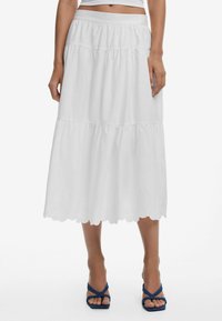 White tiered skirt with a scalloped hem; lightweight fabric with subtle floral embroidery; elastic waistband for comfort; paired with blue sandals.