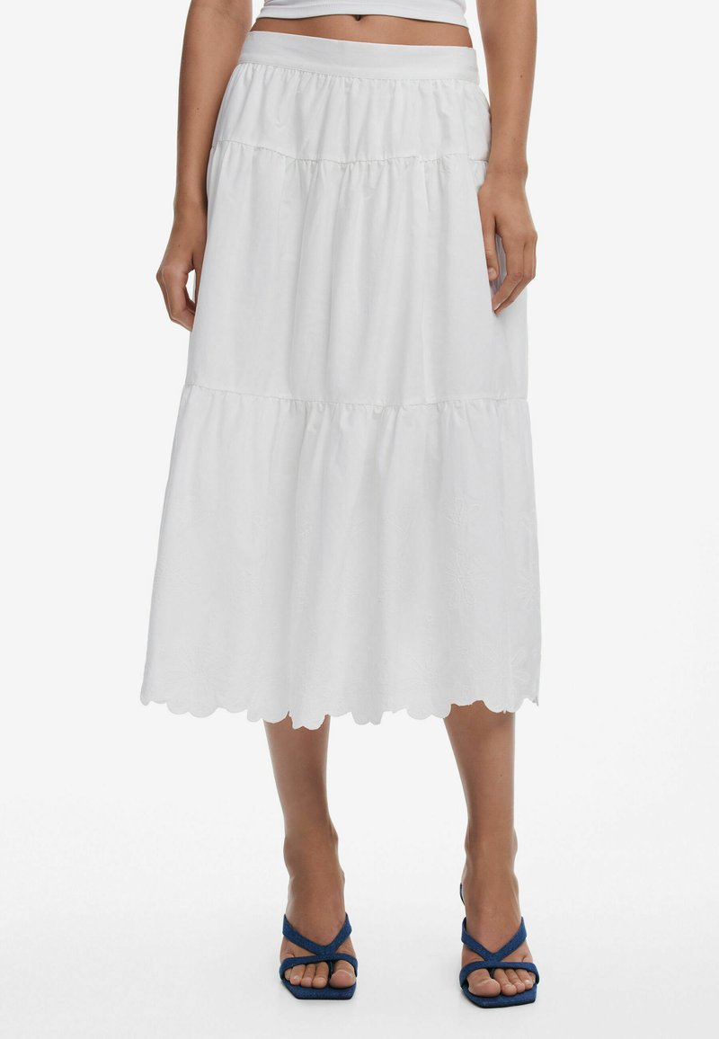 White tiered skirt with a scalloped hem; lightweight fabric with subtle floral embroidery; elastic waistband for comfort; paired with blue sandals.