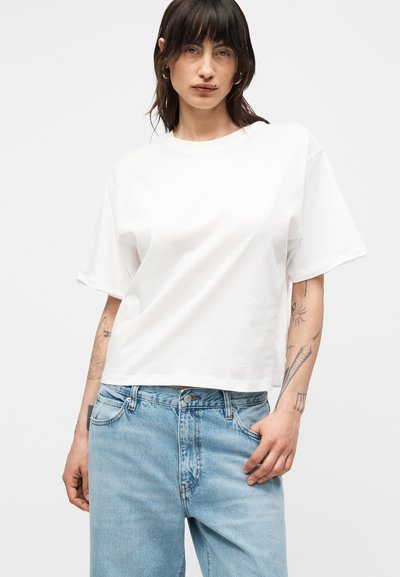 Γυναίκα με σκούρα μαλλιά, που φοράει ένα απλό λευκό υπερμεγέθες κοντομάνικο μπλουζάκι και ανοιχτό μπλε τζιν με ψηλή μέση, δείχνοντας τατουάζ στα χέρια.