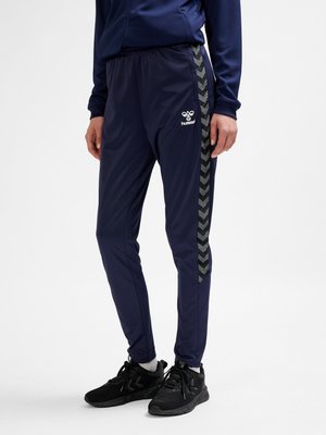 Osoba stojąca w granatowych spodniach dresowych Hummel z czarnymi paskami w jodełkę po bokach i czarnych butach sportowych.