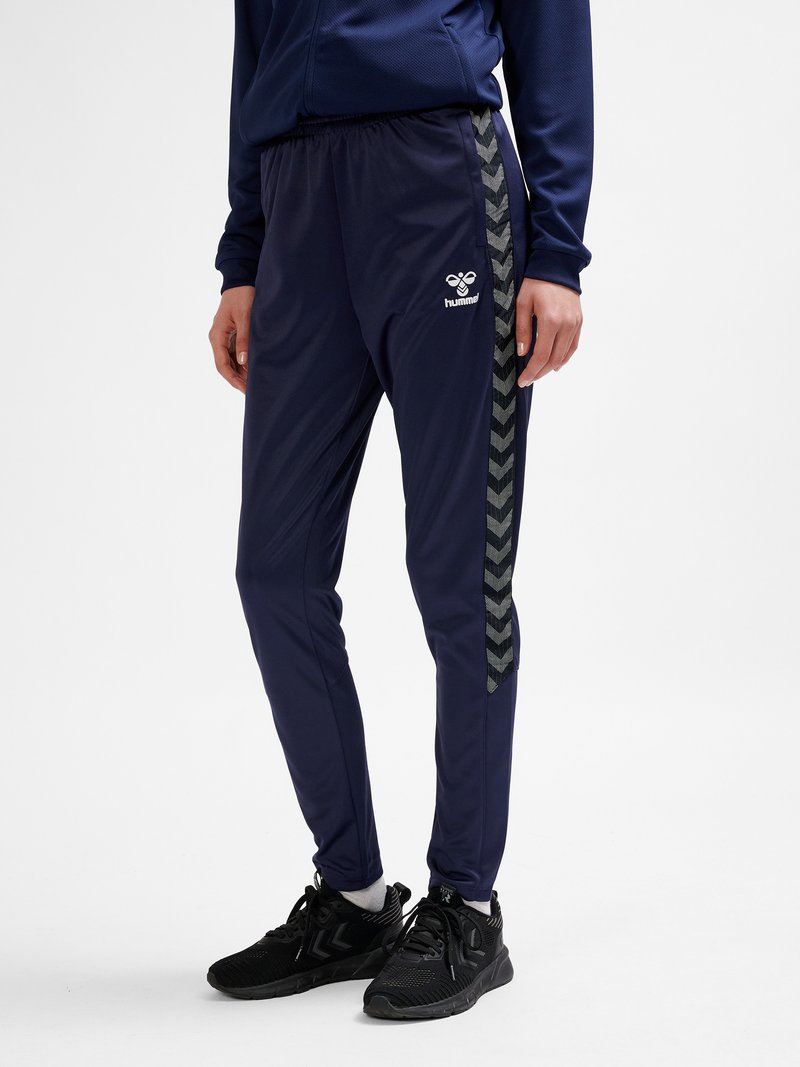 Osoba stojąca w granatowych spodniach dresowych Hummel z czarnymi paskami w jodełkę po bokach i czarnych butach sportowych.