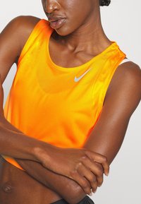 Débardeur Nike orange vif avec un col rond, un tissu léger en maille et un petit logo swoosh argenté sur la poitrine.