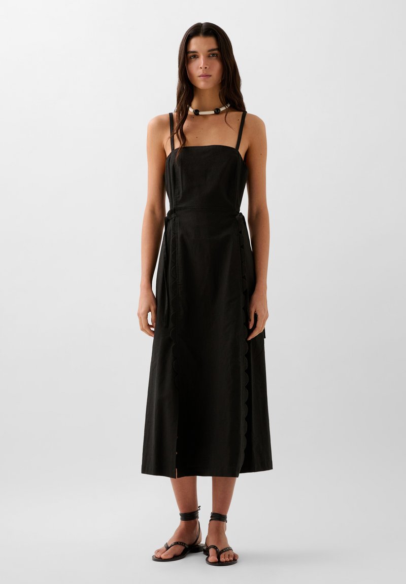 Schwarzes Kleid mit einem taillierten Oberteil und einem fließenden Rock. Mit gewelltem Detail entlang der Seite und dünnen Trägern. Kombiniert mit schwarzen Sandalen.