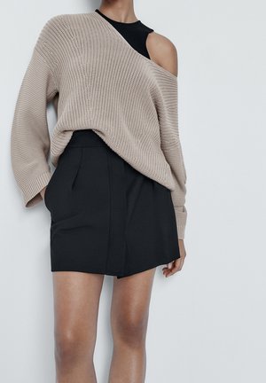 Beżowy sweter na drutach z odkrytymi ramionami, zestawiony z czarną, dopasowaną krótką spódnicą z przednim szwem i plisami.