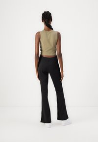 Oliwkowy krótki top na ramiączkach w połączeniu z czarnymi spodniami dzwonami i białymi trampkami, widok z tyłu na tle gładkiej białej powierzchni.