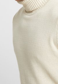 Pull beige à col roulé avec un col côtelé, fabriqué en tissu tricoté doux, présentant une texture lisse et une coupe ajustée.