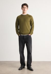 Olijfgroene lange mouw trui met geribbelde kraag, gecombineerd met donkergrijze jeans en zwarte schoenen. Model staat rechtop tegen een lichte achtergrond.