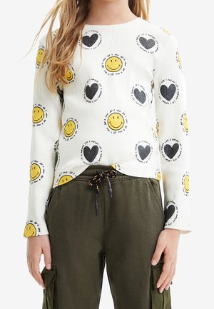 Kind draagt een wit shirt met lange mouwen met gele smileygezichten en zwarte harten, gecombineerd met olijfgroene cargobroek met een trekkoord in de taille.