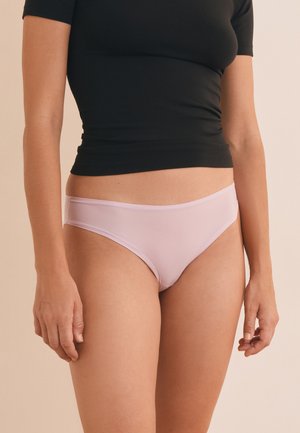 Femme portant un haut court noir et un sous-vêtement style bikini rose clair sur fond beige.