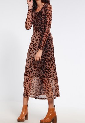 Kvinde iført en langærmet midi-kjole med leopardprint, kombineret med brune ankelstøvler, står med en hånd nær sit ansigt.