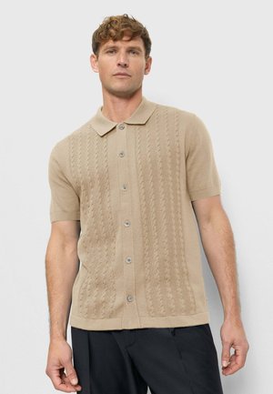 Mann mit kurzem lockigem Haar, der ein beiges, kurzärmliges, gestricktes Polohemd mit Knöpfen und eine schwarze Hose trägt, vor einem schlichten hellen Hintergrund stehend.