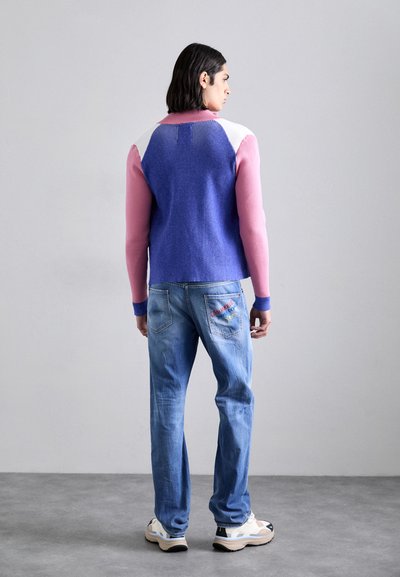 Strickpullover mit Farbblockierung in Blau, Pink und Weiß; gerippte Bündchen. Getragen mit hellblauen Jeans, die farbenfrohe gestickte Akzente aufweisen.