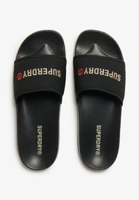 Zwarte slidesandalen met een rubberen zool, een gestructureerd oppervlak en een brede klittenbandsluiting met het "SUPERDRY" logo in wit en rood.