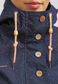 Marinblå jacka med vita prickar, träknappar, rosa dragsko och hög krage. Tillverkad av ett slätt, texturerat tyg.