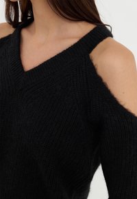 Pull noir en maille avec col en V et épaules dénudées, présentant une texture côtelée et une coupe décontractée.