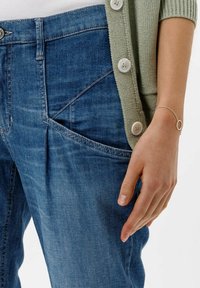 Cárdigan de punto verde claro con acentos de botones, usado sobre un jeans de mezclilla azul con un distintivo detalle de bolsillo cosido. Pulsera de plata visible.