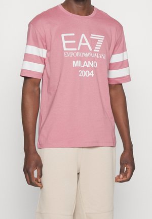 Homme portant un t-shirt rose à manches courtes avec des rayures blanches sur les manches et le texte blanc EA7 Emporio Armani Milano 2004 sur le devant.