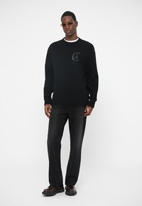 Pulover negru tricotat cu un logo texturat, purtat cu blugi negri și pantofi lucioși. Modelul stă în fața unui fundal alb simplu.