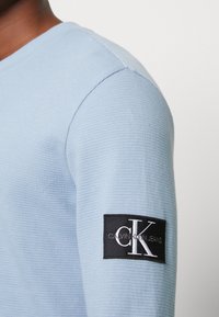 Světle modrá texturovaná košile s dlouhým rukávem a černou obdélníkovou nášivkou loga Calvin Klein Jeans na horním rukávu.