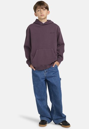 Paarse hoodie met een voorzak en klein logo, gecombineerd met losse blauwe denim jeans en zwarte sneakers met rubberen zolen.