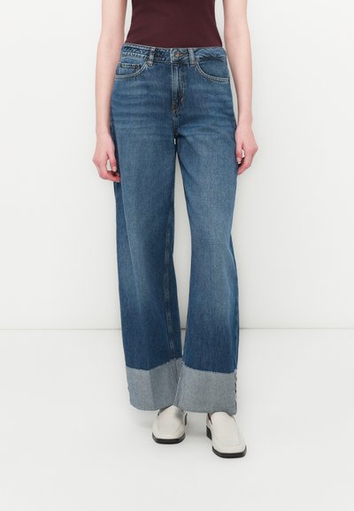 Jeans en denim bleu à jambes larges avec un ourlet effiloché, dotés d'une taille haute et de cinq poches, accompagnés d'un haut sombre et de chaussures claires.