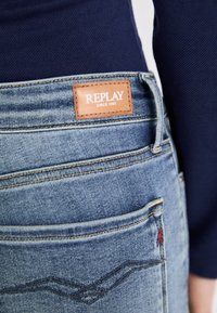 Blue denim jeans with a brown leather label reading "REPLAY SINCE 1981." Features contrast stitching, a classic pocket design, and slight fading.