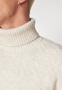 Maglione bianco a collo alto lavorato a maglia, con un colletto a coste e motivi a zigzag testurizzati, in primo piano su colletto e spalla.