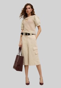 Pantaloni cargo beige con tasche laterali, abbinati a un top lavorato a maglia con maniche a palloncino. Gli accessori includono una cintura nera e una borsa a spalla bordeaux.