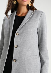 Blazer gris en tissu texturé, coupe croisée avec trois boutons. Comprend une poche poitrine et des revers crantés.