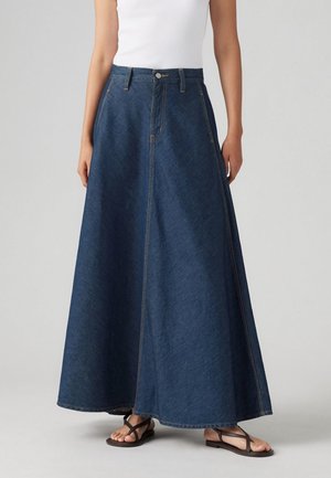 Femme portant une longue jupe trapèze en denim bleu foncé avec des poches, associée à des sandales plates noires à brides et un top blanc sans manches.