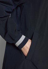 Námořnická bunda s strukturovanou tkaninou; má elastické manžety s šedými a námořnickými pruhy. Obsahuje boční kapsu a elegantní, minimalistický design.