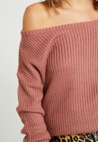 Femme portant un pull en maille rose poussiéreux à épaules dénudées et un pantalon imprimé léopard avec une boucle de ceinture visible sur un fond uni.