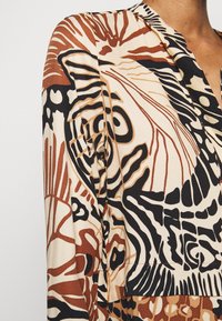Top in tessuto stampato con tonalità di marrone, nero e beige, caratterizzato da disegni astratti e superficie testurizzata. Collo a V e dettaglio con bottoni.