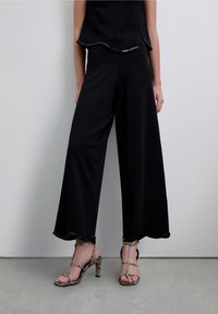 Pantalones negros de pierna ancha con un dobladillo con borde ondulado, combinados con sandalias de tacón alto con estampado de piel de serpiente y múltiples tiras.