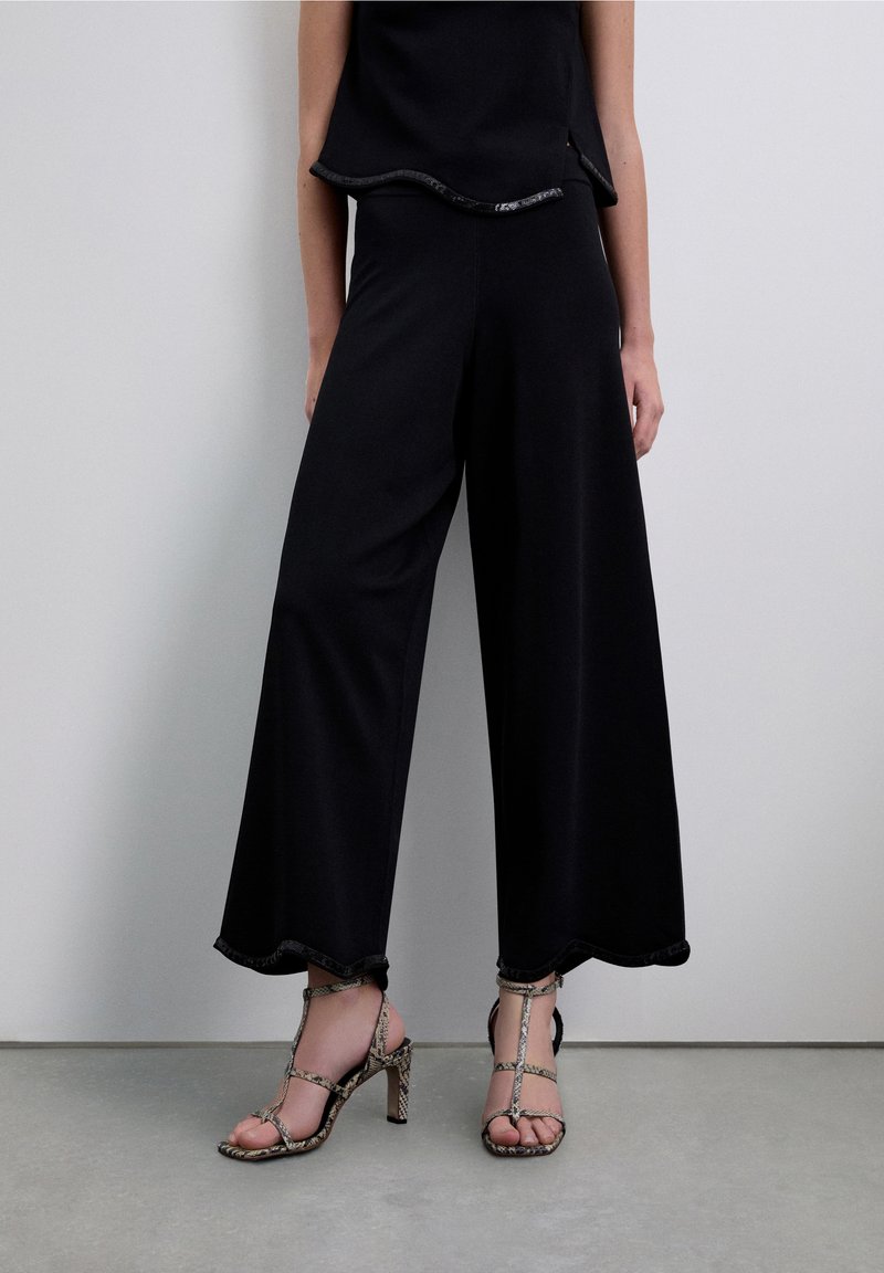 Pantalones negros de pierna ancha con un dobladillo con borde ondulado, combinados con sandalias de tacón alto con estampado de piel de serpiente y múltiples tiras.