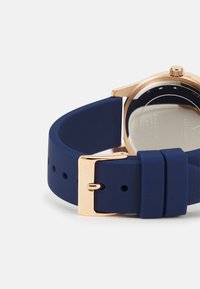Correa de reloj de silicona azul marino con hebilla metálica dorada; la parte trasera del reloj presenta una superficie metálica suave con grabados.