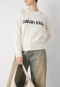 Hvit strikket genser med svart "calvin klein" tekst; rillede mansjetter og hem. Modellen holder en beige lærveske; blå denim bukser.