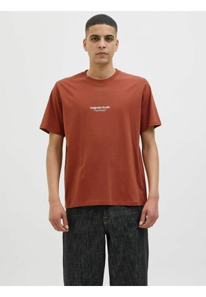 Rust-colored cotton t-shirt with short sleeves, round neckline, and centered white "Originals Studio" text logo. Worn with dark jeans.