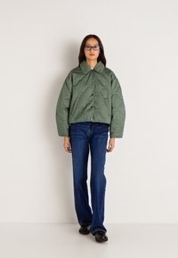 Πρασίνου επενδεδυμένο μπουφάν με φαρδύ γιακά και μπροστινά κουμπιά, συνδυασμένο με μπλε φαρδύ τζιν και μαύρα παπούτσια. Απλή υπόθεση.