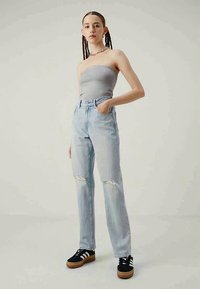 Γυναίκα με πλεξούδα, ντυμένη με αμάνικο γκρι τοπ, ανοιχτόχρωμα σκισμένα τζιν και μαύρα αθλητικά παπούτσια με λευκές ρίγες, που στέκεται με το ένα χέρι στην τσέπη.