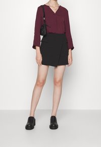 Haut bordeaux à manches longues avec un décolleté en V et une jupe portefeuille noire avec des poches avant, assorti à des chaussures à talons carrés noires.