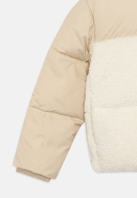 Giacca imbottita beige con un morbido pannello anteriore in shearling bianco. Presenta un design trapuntato e polsini a costine per calore e comfort.