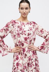 Γυναίκα που φοράει λευκό φόρεμα με μακριά μανίκια και ροζ ανθισμένο σχέδιο, με χρυσή ζώνη αλυσίδα, τα χέρια της στη μέση, κοιτάζοντας προς τα πλάγια.