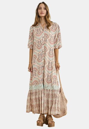 Mujer con un vestido largo abotonado con estampado y mangas abullonadas, sosteniendo un bolso tejido y llevando sandalias de plataforma.