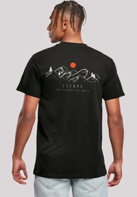 Schwarzes Baumwoll-T-Shirt mit einem Berg-Graphic, roter Sonne und weißem Text "ENTFLIEH UND ENTDECKE DIE WELT" auf dem Rücken. Kurzärmlig.
