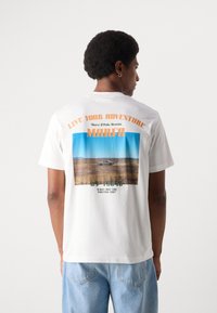 Weißes Baumwoll-T-Shirt mit grafischem Druck auf dem Rücken, der eine Wüstenlandschaft zeigt, Text in Orange und Grün sowie einem runden Halsausschnitt.