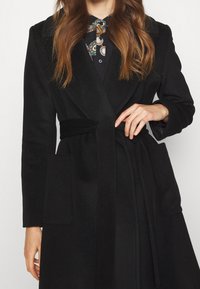 Svart ullblandad kappa med sjalkrage, bälte i midjan och två framfickor. Slät textur och skräddarsydd passform, lämplig för lager-på-lager.